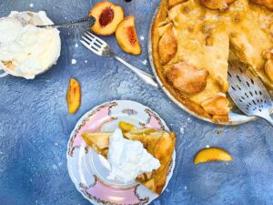 An overhead view of a pie with a slice out of it. There is a pink floral plate with a slice of pie on it. There are sliced peaches and cream in the background.