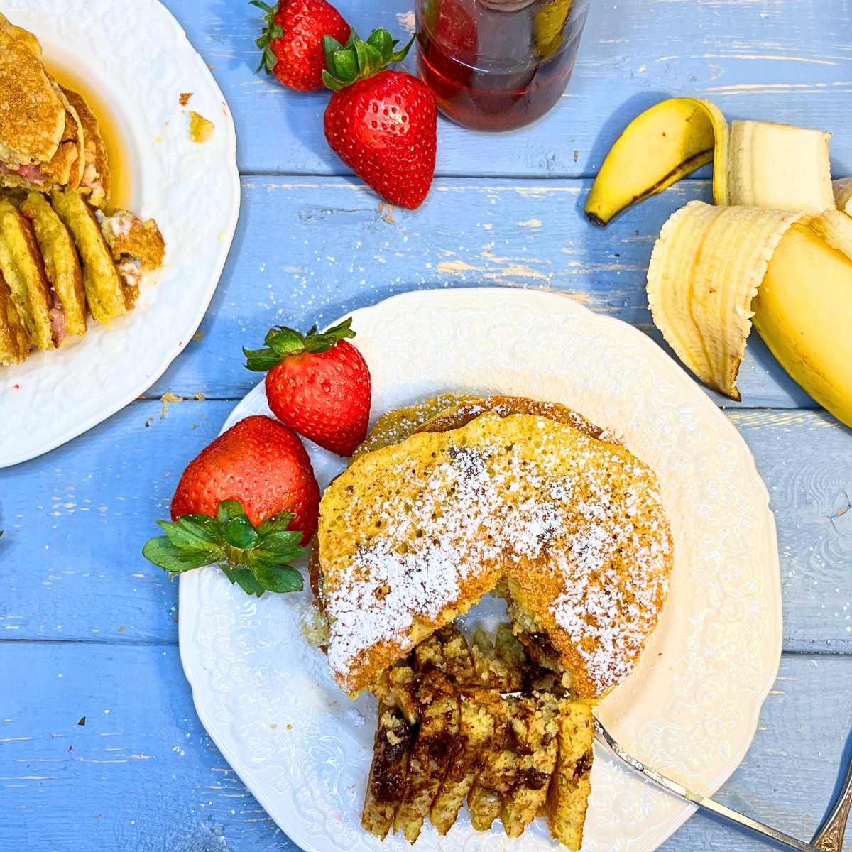 There is a blue table with pancakes with a slice out of them. There is powdered sugar on top. There are strawberries and bananas.