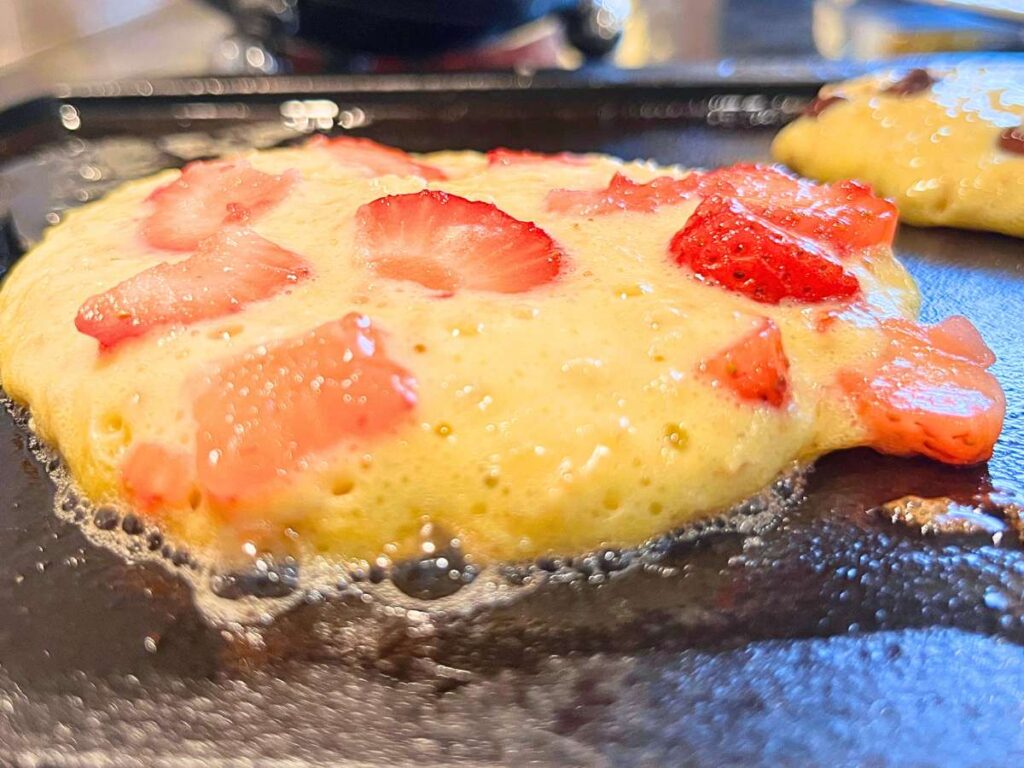 There are pancakes cooking on a griddle with strawberries inside.