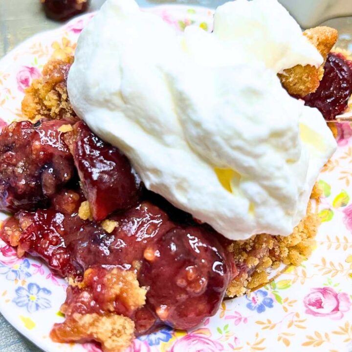 A close up of a cherry crumble on a floral plate with whipped cream on top.