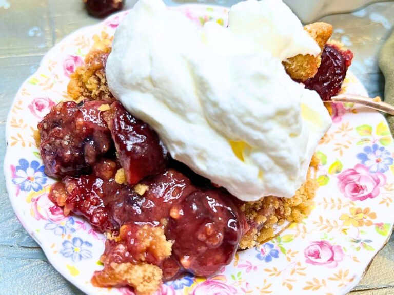 A close up of a cherry crumble on a floral plate with whipped cream on top.