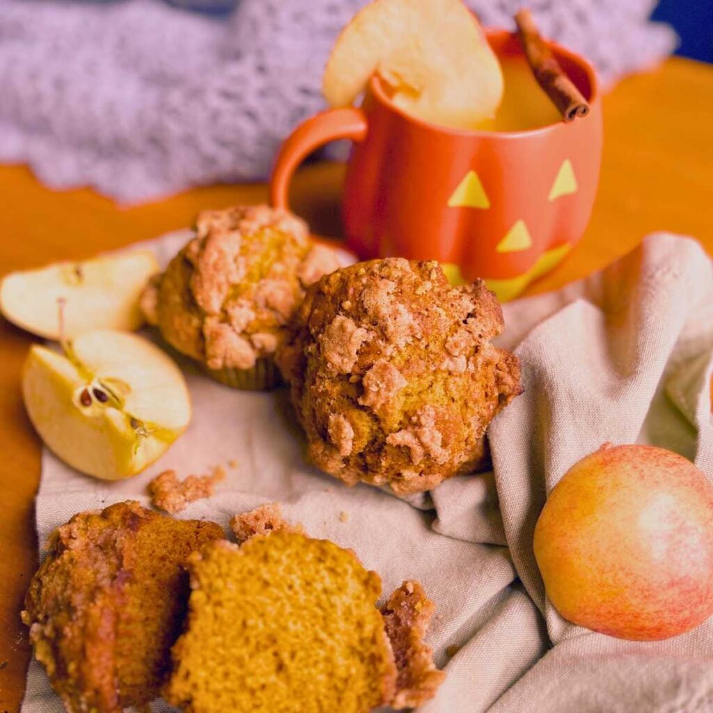 There are some pumpkin muffins on a wooden table. There is an apple and a pumpkin mug in the background.