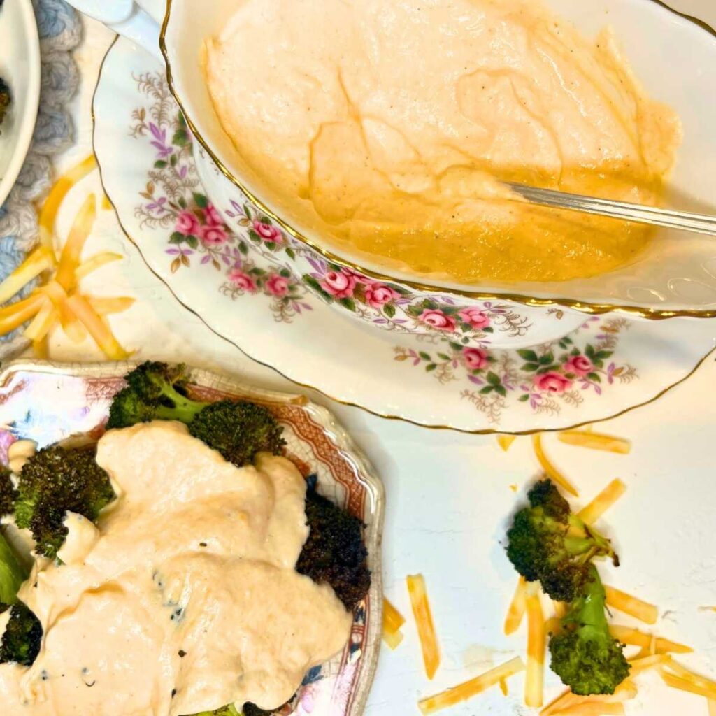 There is a bowl of cheese sauce with a spoon in it. There is a plate of broccoli in front with cheese sauce on top.