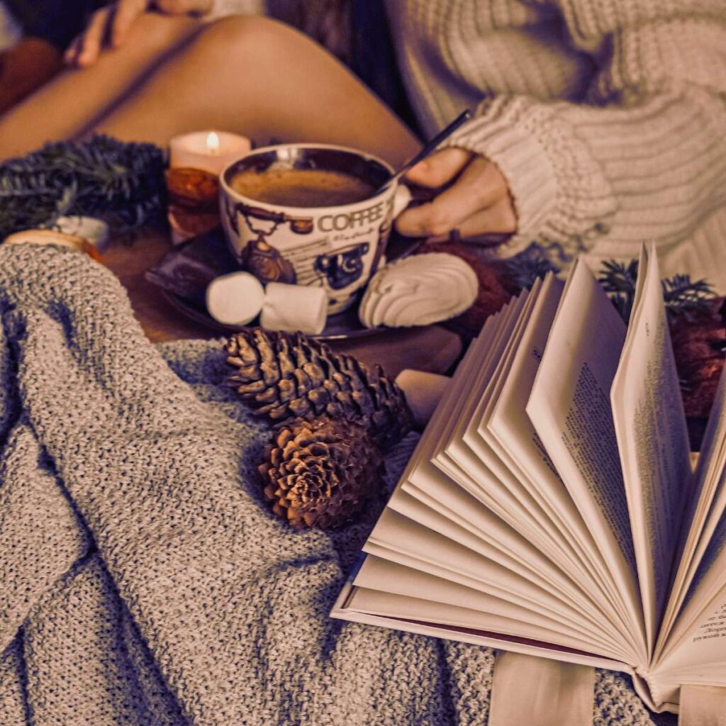 There is a woman sitting in blankets with a cup of hot chocolate. There is a book in from and some pinecones.