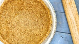 A baked graham cracker crust in a metal pie plate. There is a rolling pin beside.