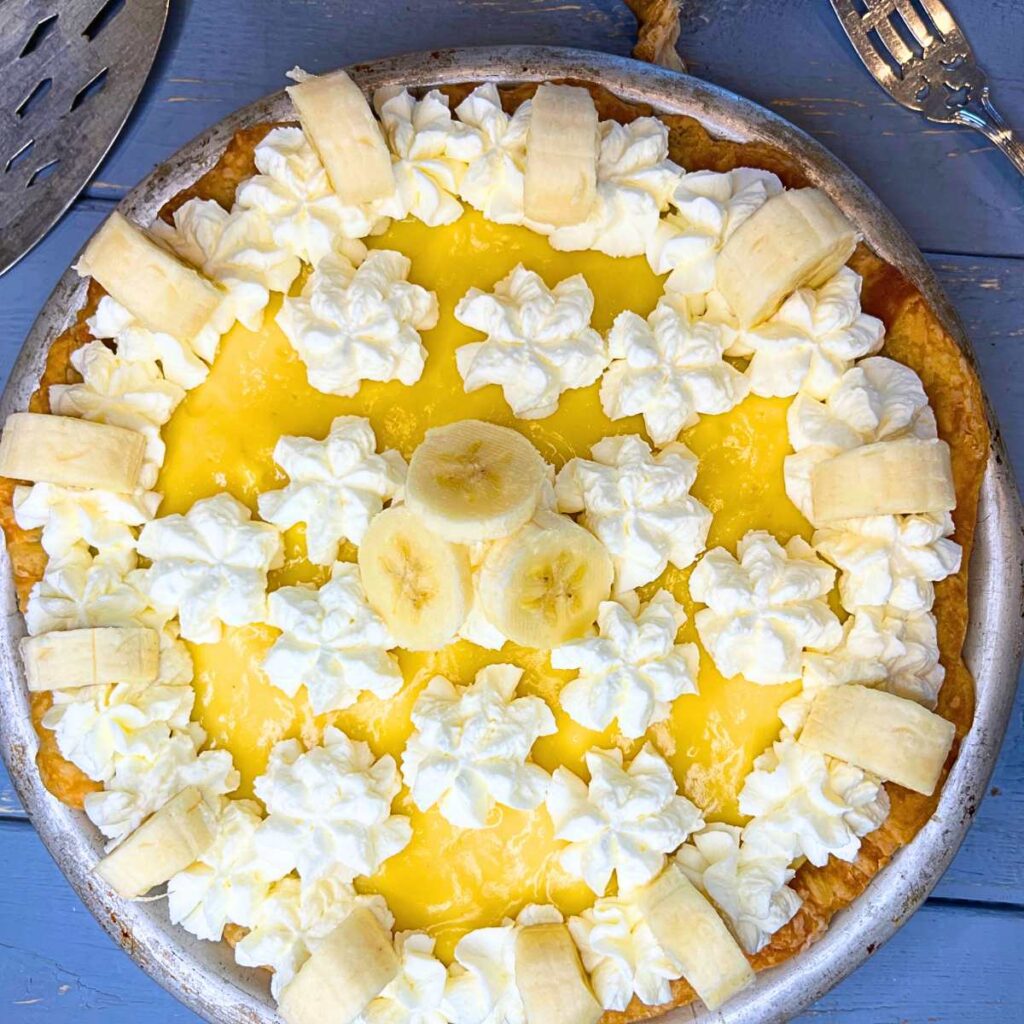 A banana cream pie on a blue table decorated with whipped cream and fresh bananas.