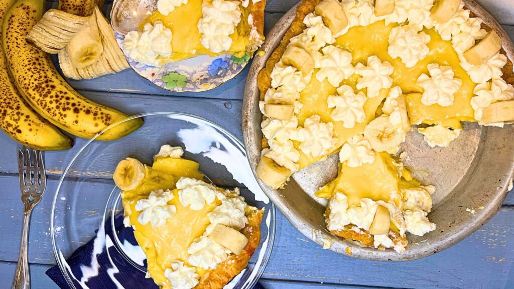 A sliced banana cream pie on a blue table. There is a slice of pie on a glass plate and another one behind.