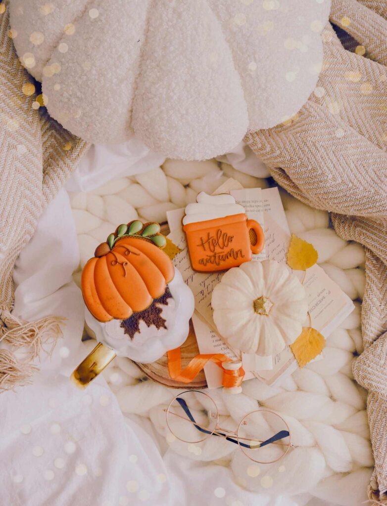 There is a collection of textured white blankets. There is a coffee and some decorated fall cookies on top. There are a few white pumpkins around.