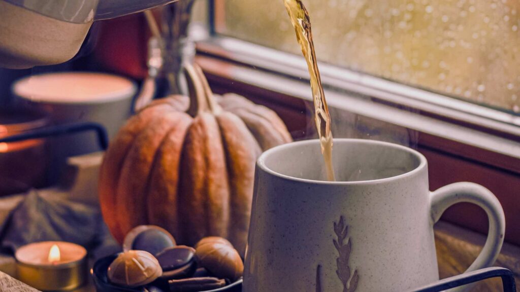 There is tea being poured into a cup. behind is pumpkins and acorns.