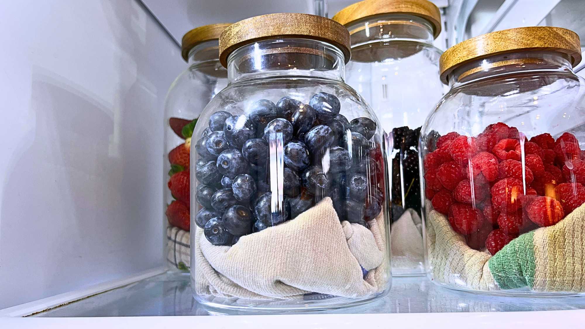 There are some berries in glass jars in a refrigerator.