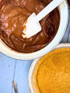 There is a pot of chocolate filling with a white spatula inside. There is a graham cracker crust in front.