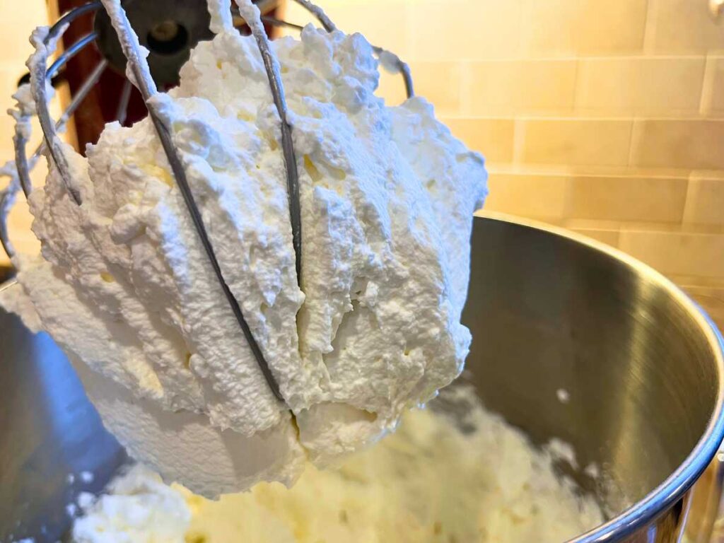 The whisk attachment for a stand mixer full of whipped cream. There is more in the bowl in the background.