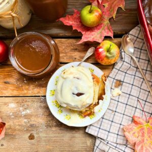 There is a cinnamon roll with white icing on a wooden table. There is a jar of apple butter beside.