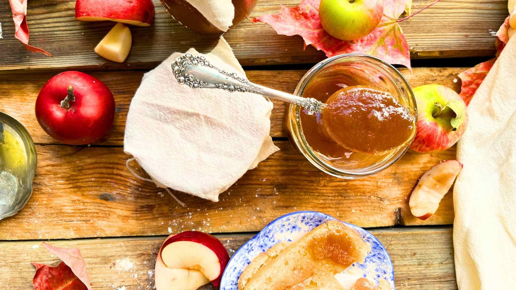 An open jar of apple butter with a spoon resting on top. There is a sealed jar beside. There are apples around.