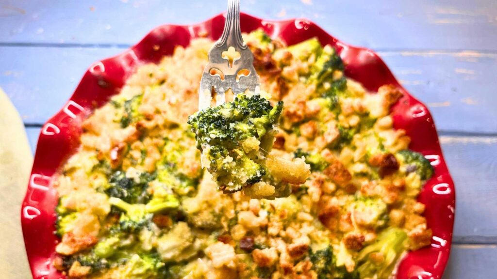 There is a broccoli casserole in a red dish. A fork is holding up a spear of broccoli.