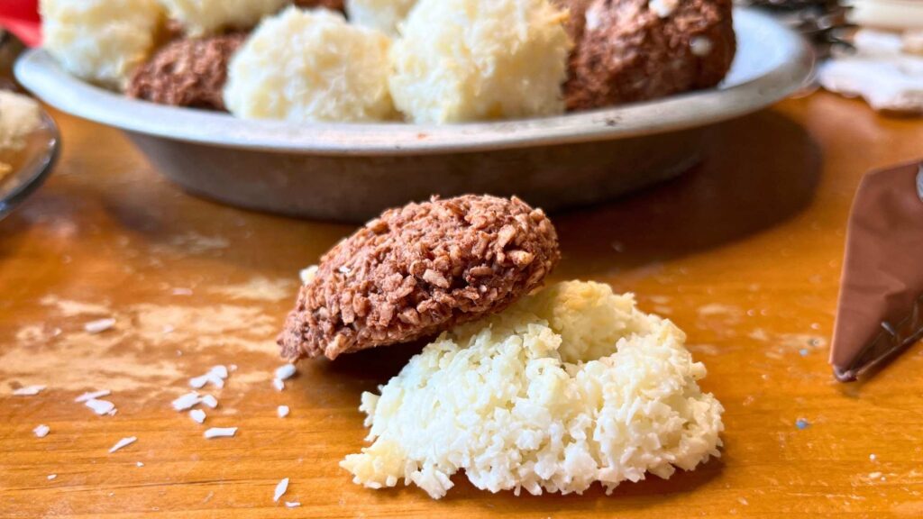 There is one chocolate and one coconut macaroon on a wooden table. There are more macaroons in the background.