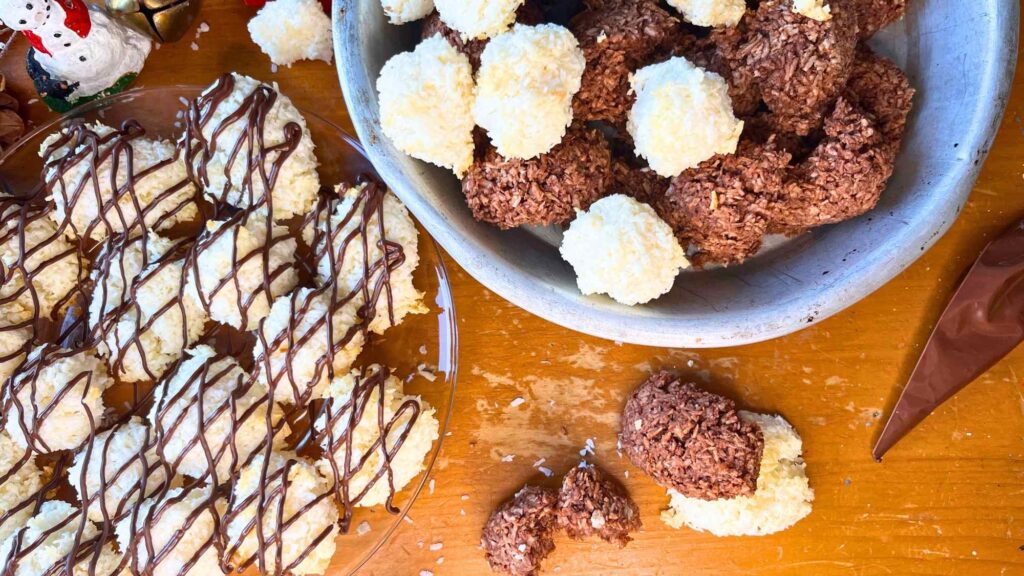 A glass plate of coconut macaroons drizzled with chocolate. There is a metal pie plate with coconut and chocolate macaroons inside.