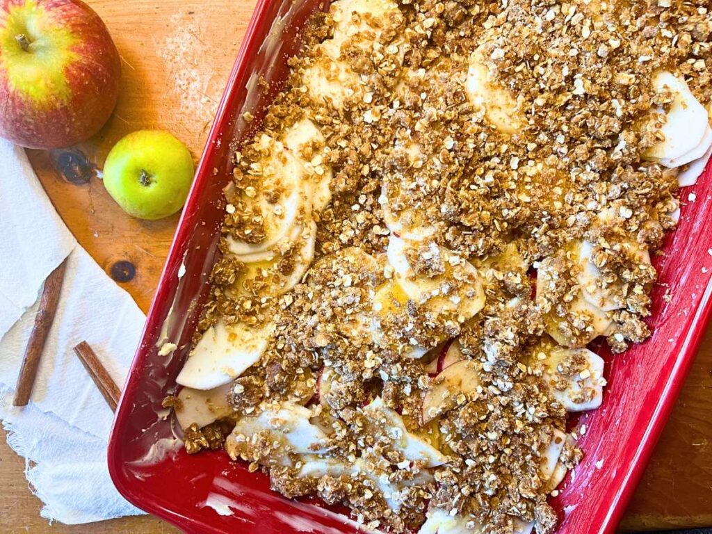 A red casserole dish with sliced apples, honey, and a oat crumble topping inside.