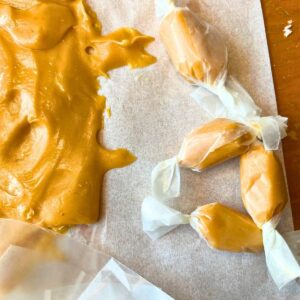 Some wrapped caramel candies beside a big batch of caramel on parchment paper.