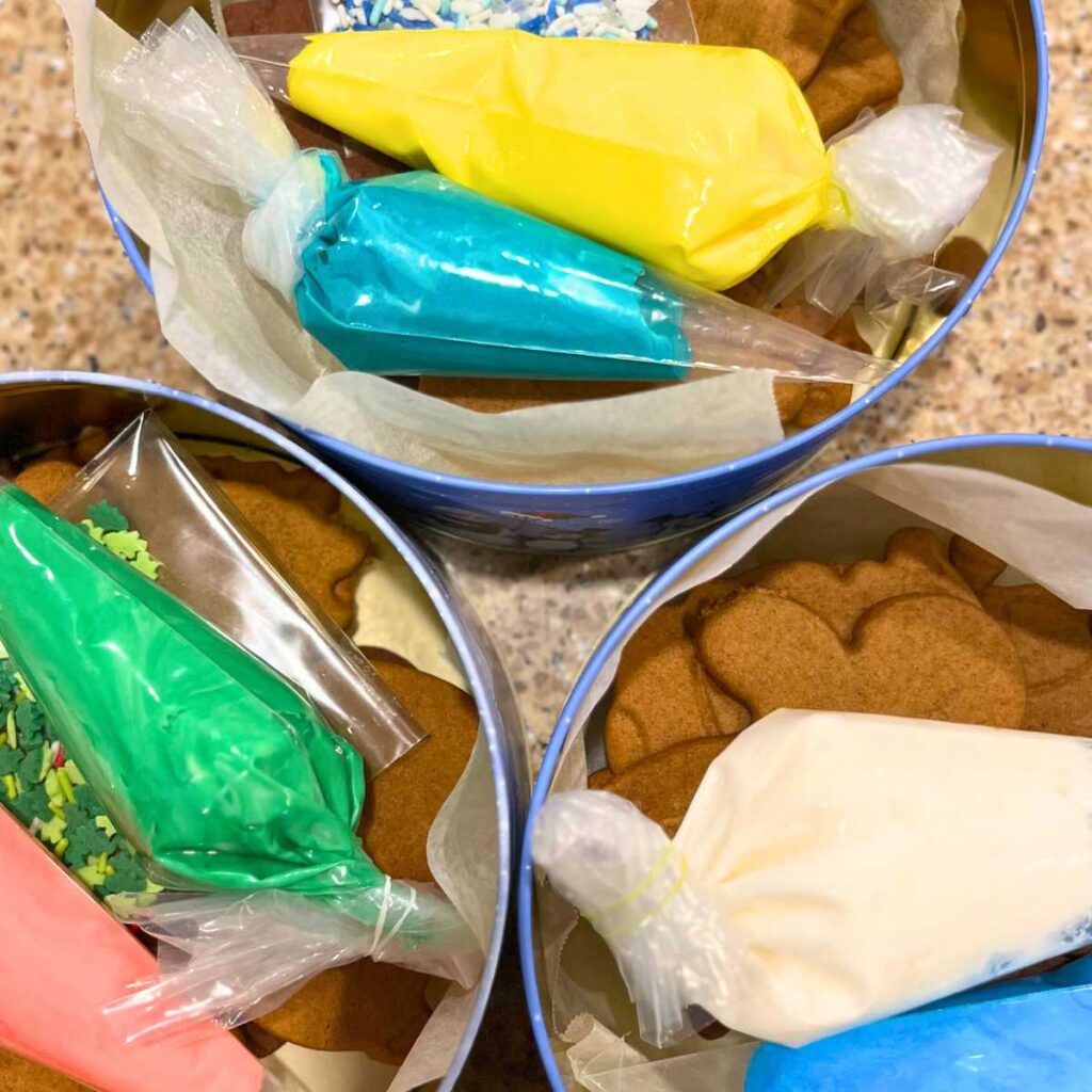 Three Christmas tins with gingerbread cookies, piping bags of coloured icing, and sprinkles inside.