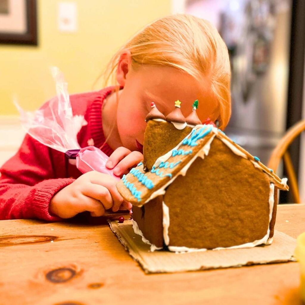 A girl is decorating a gingerbread house with a pink icing bag.