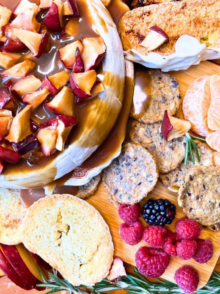 A close up look at a baked Brie cheese covered in caramel and apple pieces. There are crackers and fresh fruit around.