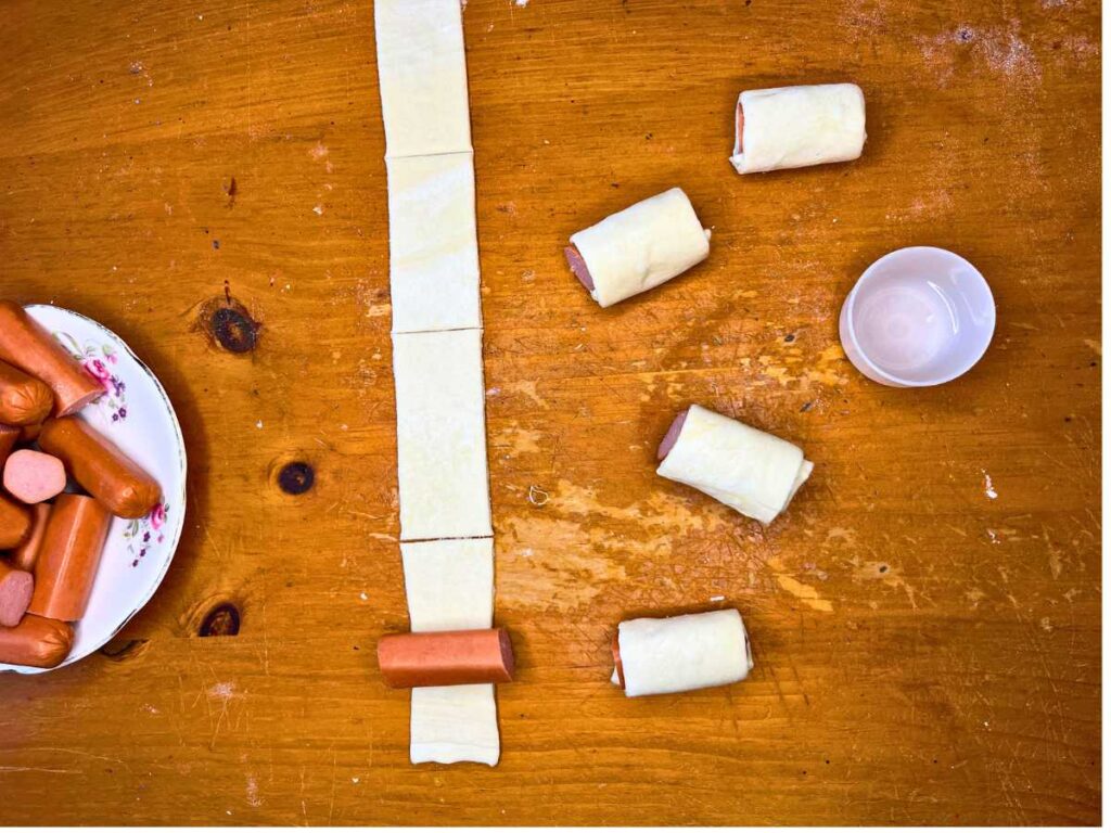 Strips of puff pastry cut into long rectangles. There are sliced hotdogs getting rolled inside.