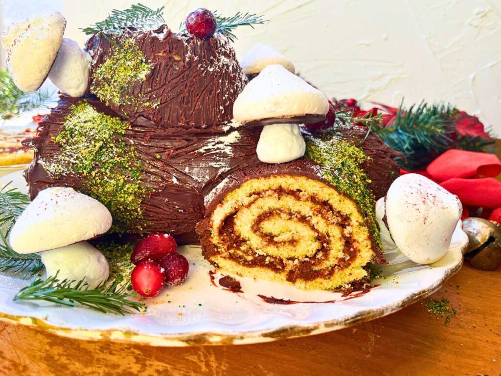 A chocolate cake in the shape of a log. It is decorated with meringue mushrooms, cranberries, and evergreen tree pieces.