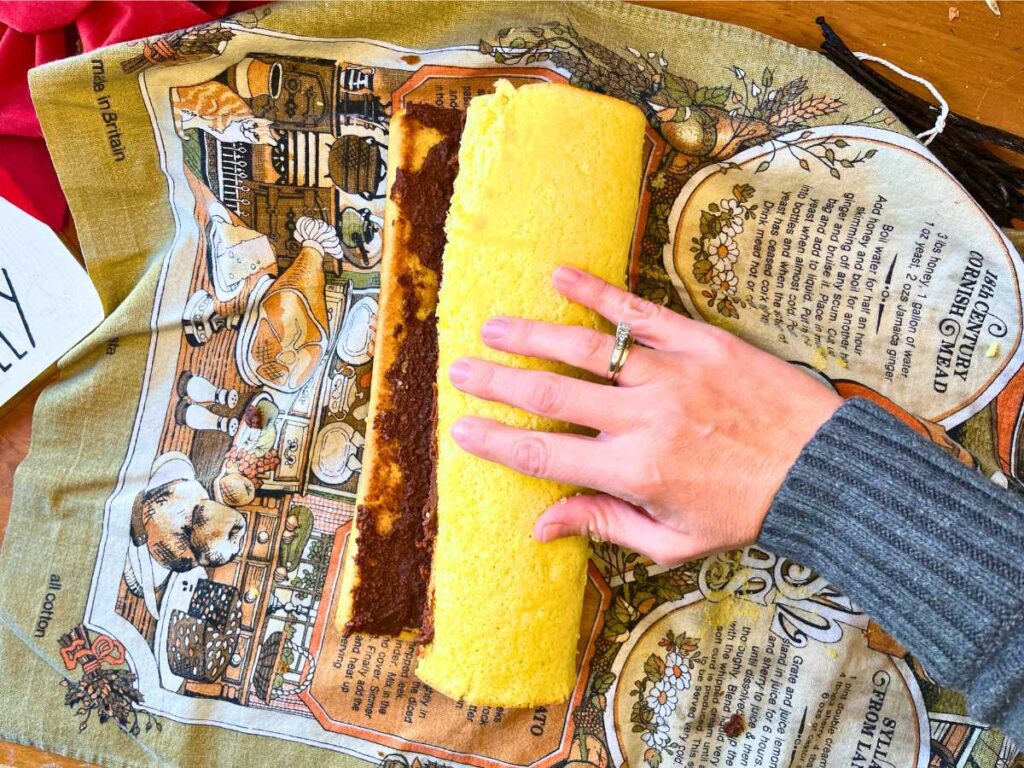 A woman is rolling a vanilla jelly roll with chocolate icing inside.