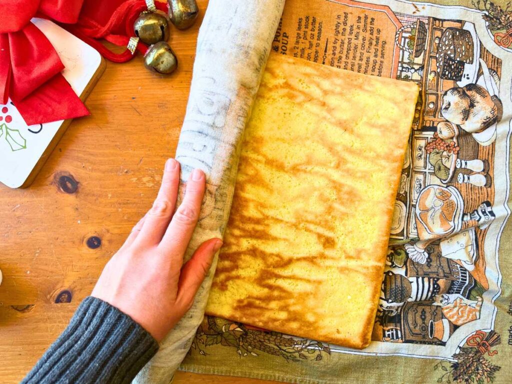 A woman rolling a rectangle of vanilla cake into a spiral with a kitchen towel.