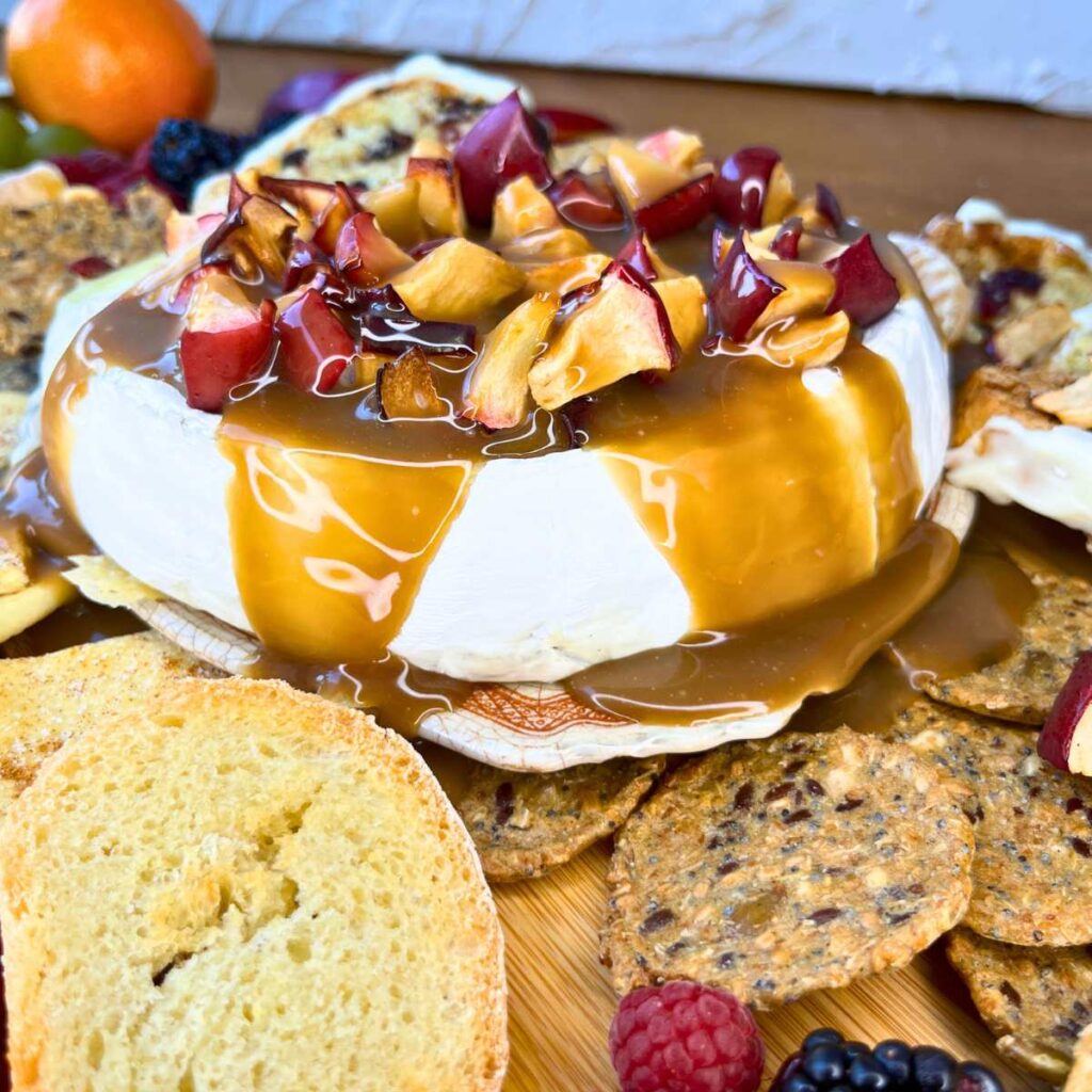 A baked Brie covered in caramel and apple pieces. Surrounded by crackers and fresh fruit.