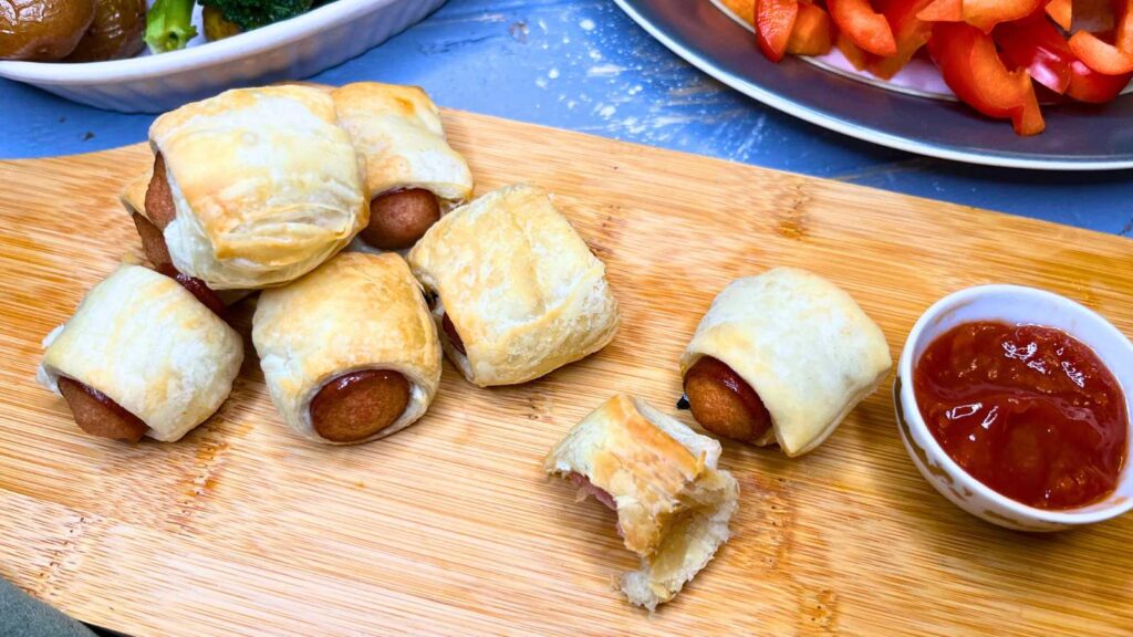 Small hotdogs wrapped in puff pastry on a wooden serving board. One has a bite out of it.