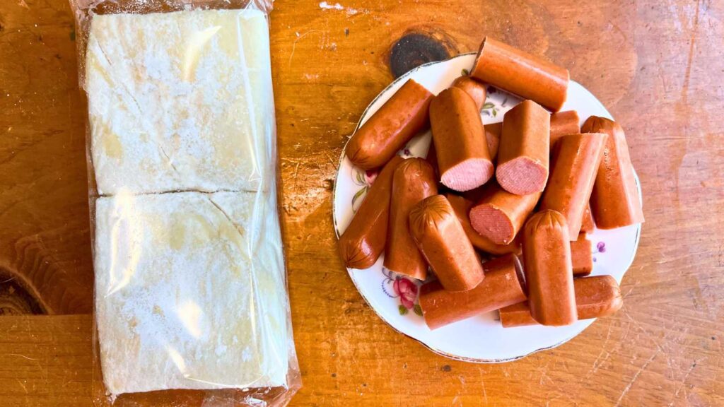 A package of puff pastry dough and a plate of sliced hotdogs on a wooden table