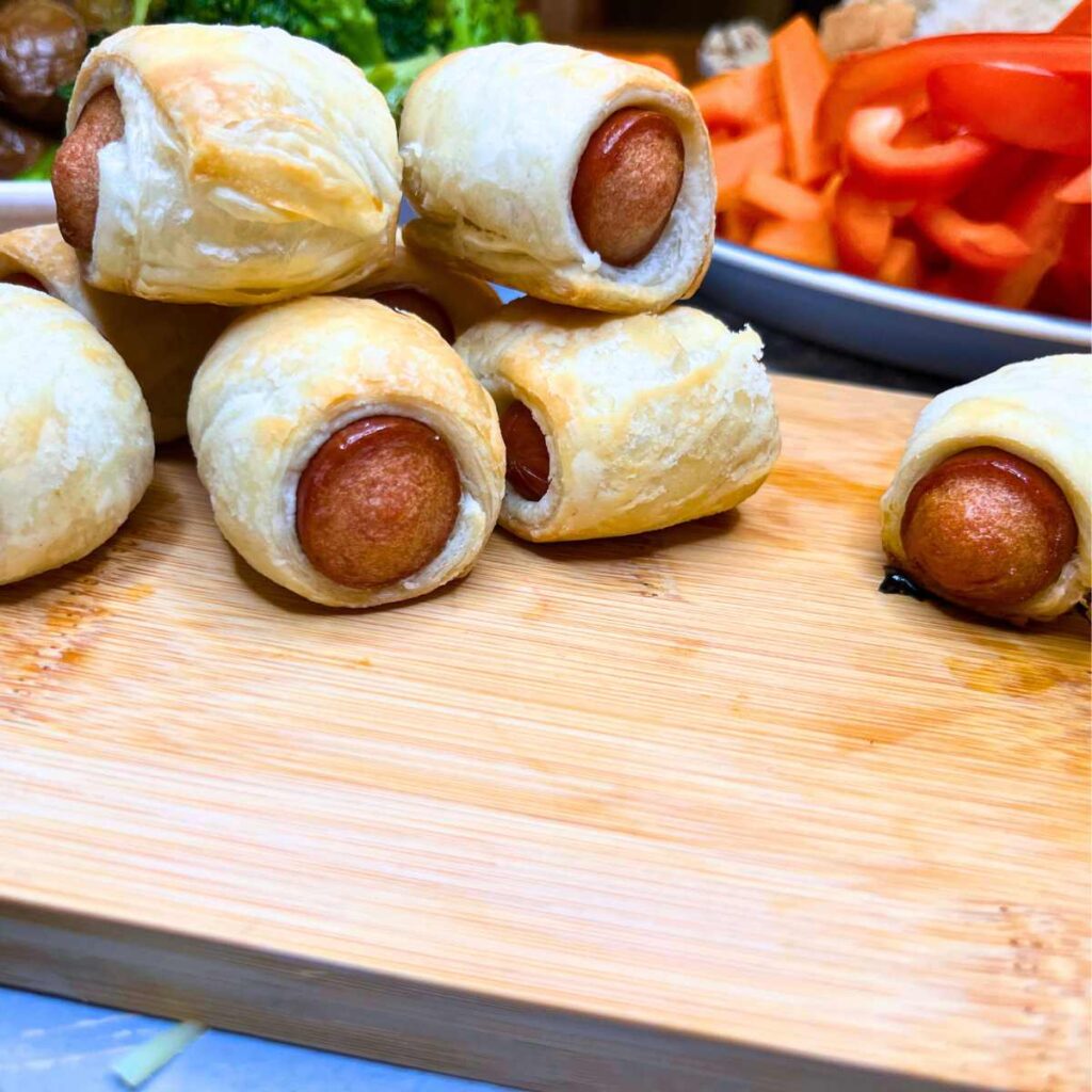 A stack of puff pastry wrapped around hotdogs. There is sliced fresh vegetables in the background.