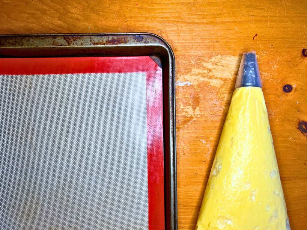 A lined baking sheet with a filled pastry bag beside. It is on a wooden table.