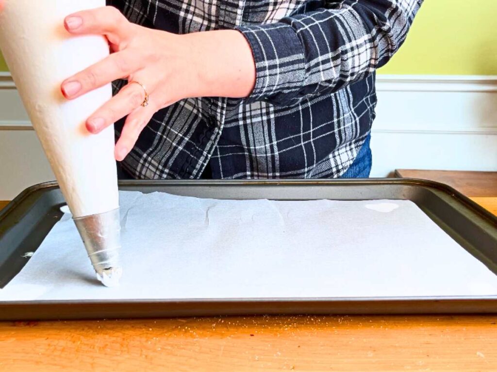 A woman is holding a piping bag full of white meringue. There is a baking sheet lined with parchment paper in front of her.