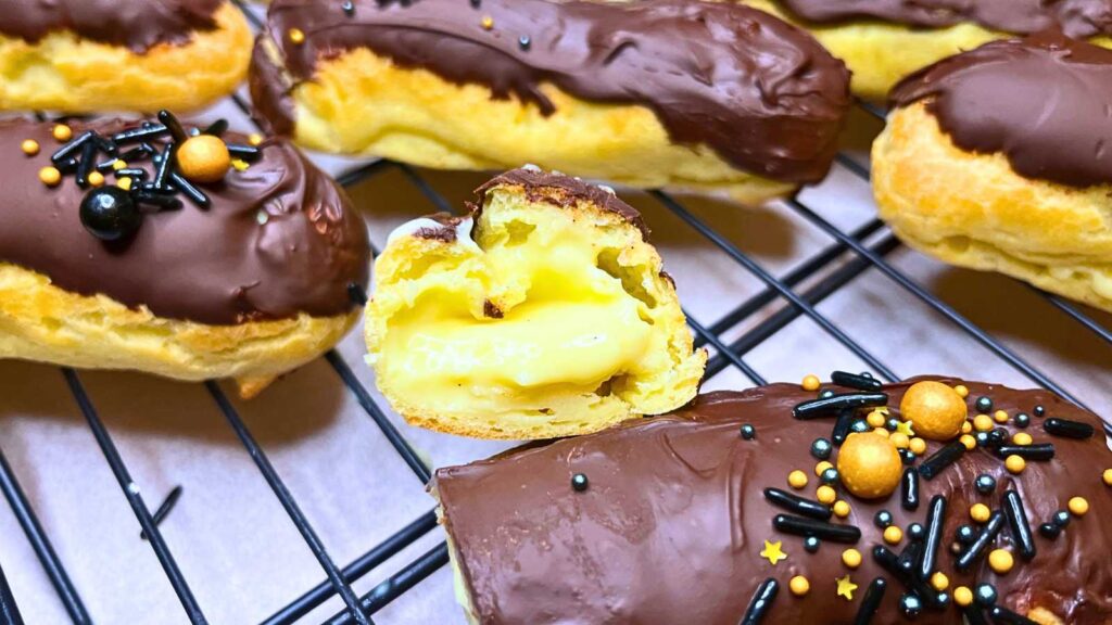 Eclairs dipped in chocolate on a wire rack. There are sprinkles on top. One of the eclairs is cut in half to. show the pastry cream in side.