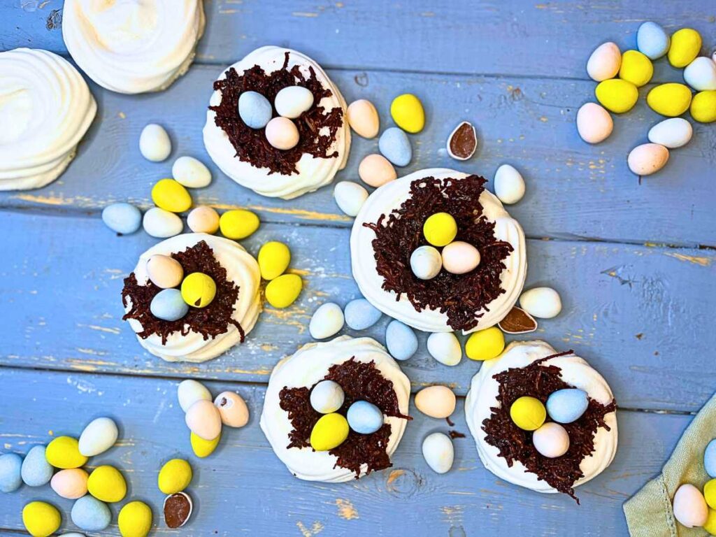 Five easter meringue nests filled with chocolate and mini candy eggs. There are more eggs around.