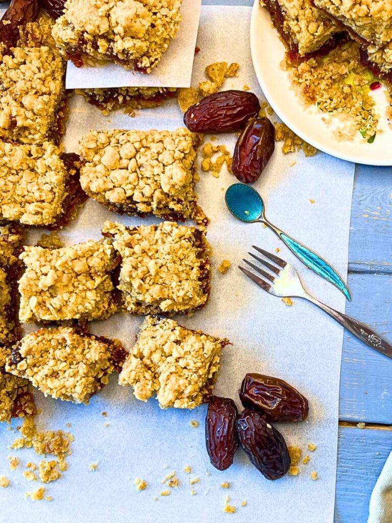 Date squares on a piece of parchment paper. There are whole dates and a plate of date squares to the side.