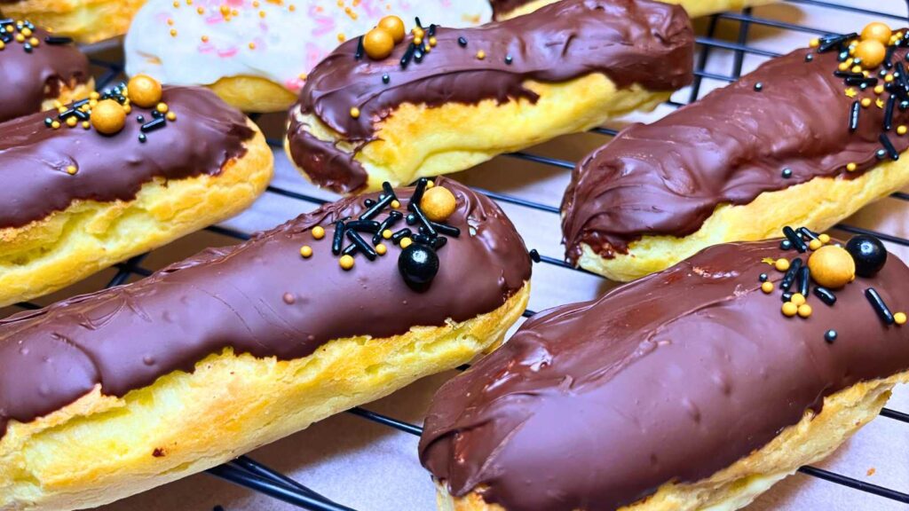 A group of chocolate eclairs on a wire rack. They are decorated with sprinkles.