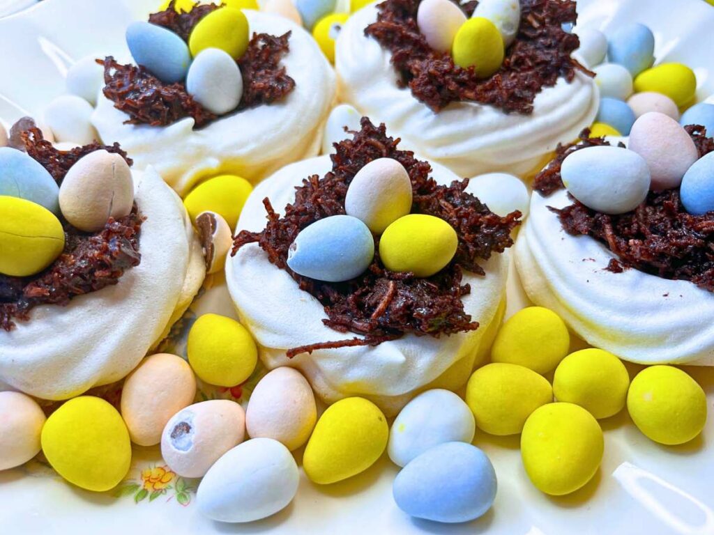 A group of meringue nests filled with chocolate and mini candy eggs. There are more eggs arranged around.