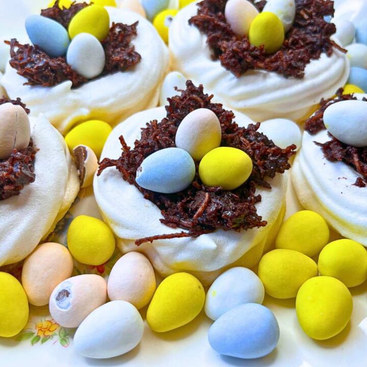 A group of meringue nests filled with chocolate and mini candy eggs. There are more eggs arranged around.