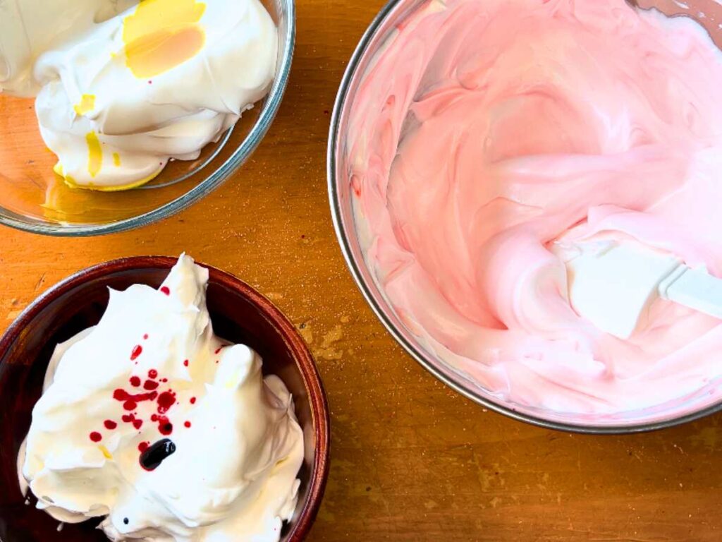 There are three bowls with whipped French meringue. One is filled with pink meringue. The other two have meringue with some drips of red and yellow food colouring on top.