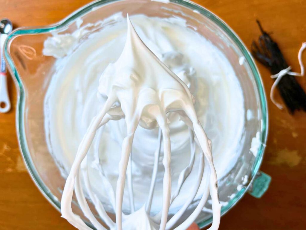 A woman is holding up a whisk attachment showing meringue with a stiff peak. There is more meringue in the bowl behind.