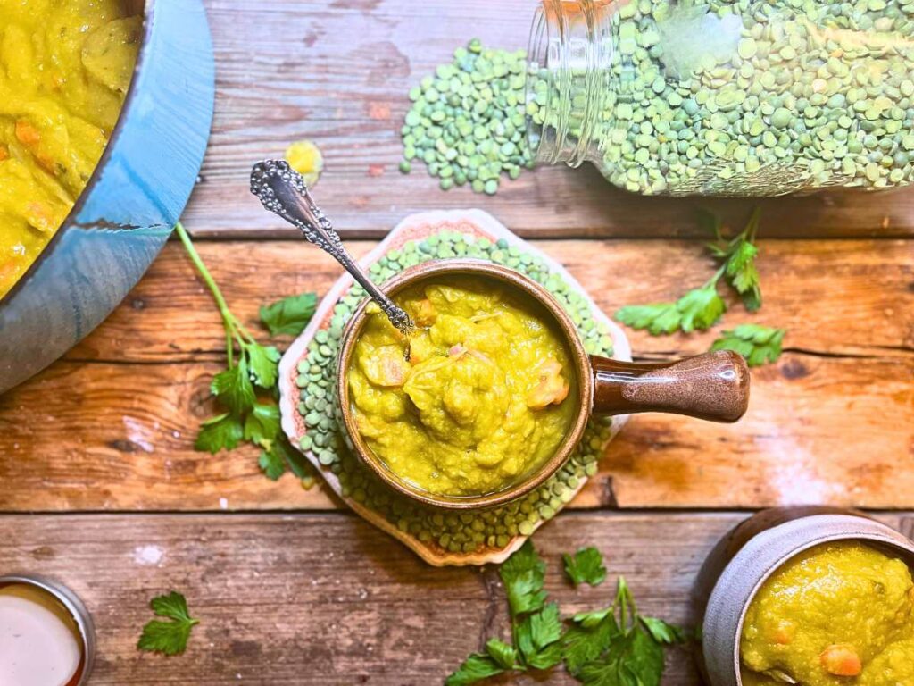 An overhead look at a bowl of pea soup with a spoon inside. There is another bowl off to the side and a pot with more of the soup.