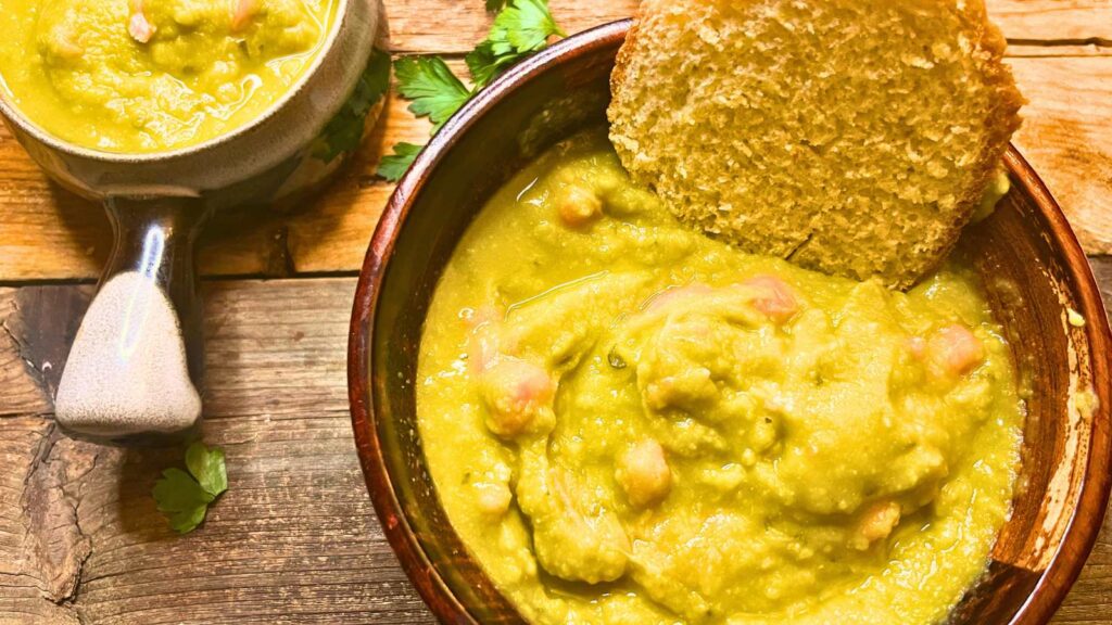 Two bowls of split pea soup on a wooden table. One bowl has a slice of bread in it.