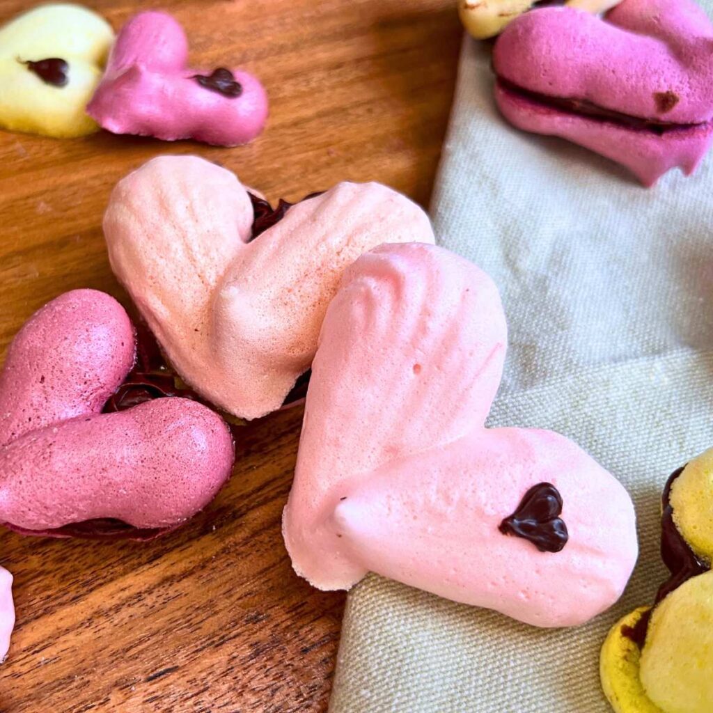 A collection of heart shaped French meringue cookies. Some are decorated with small chocolate hearts. Some are sandwiched with chocolate ganache.