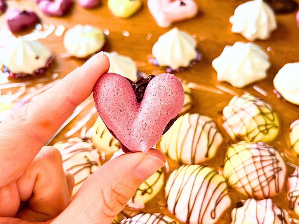 A woman is holding a purple heart meringue sandwiched with chocolate ganache.