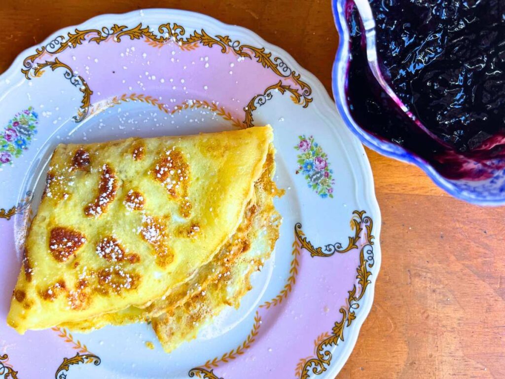 A pink and white plate with a folded crêpe. There is blueberry sauce off to the side.