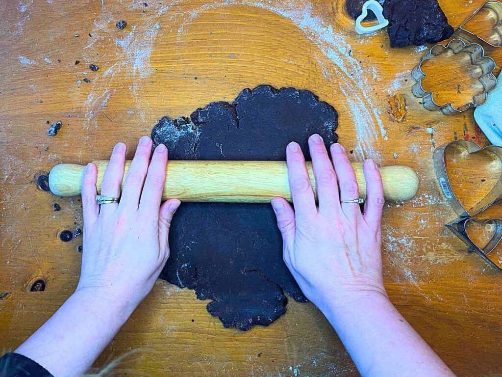 A woman is rolling out chocolate cookie dough with a wooden rolling pin.